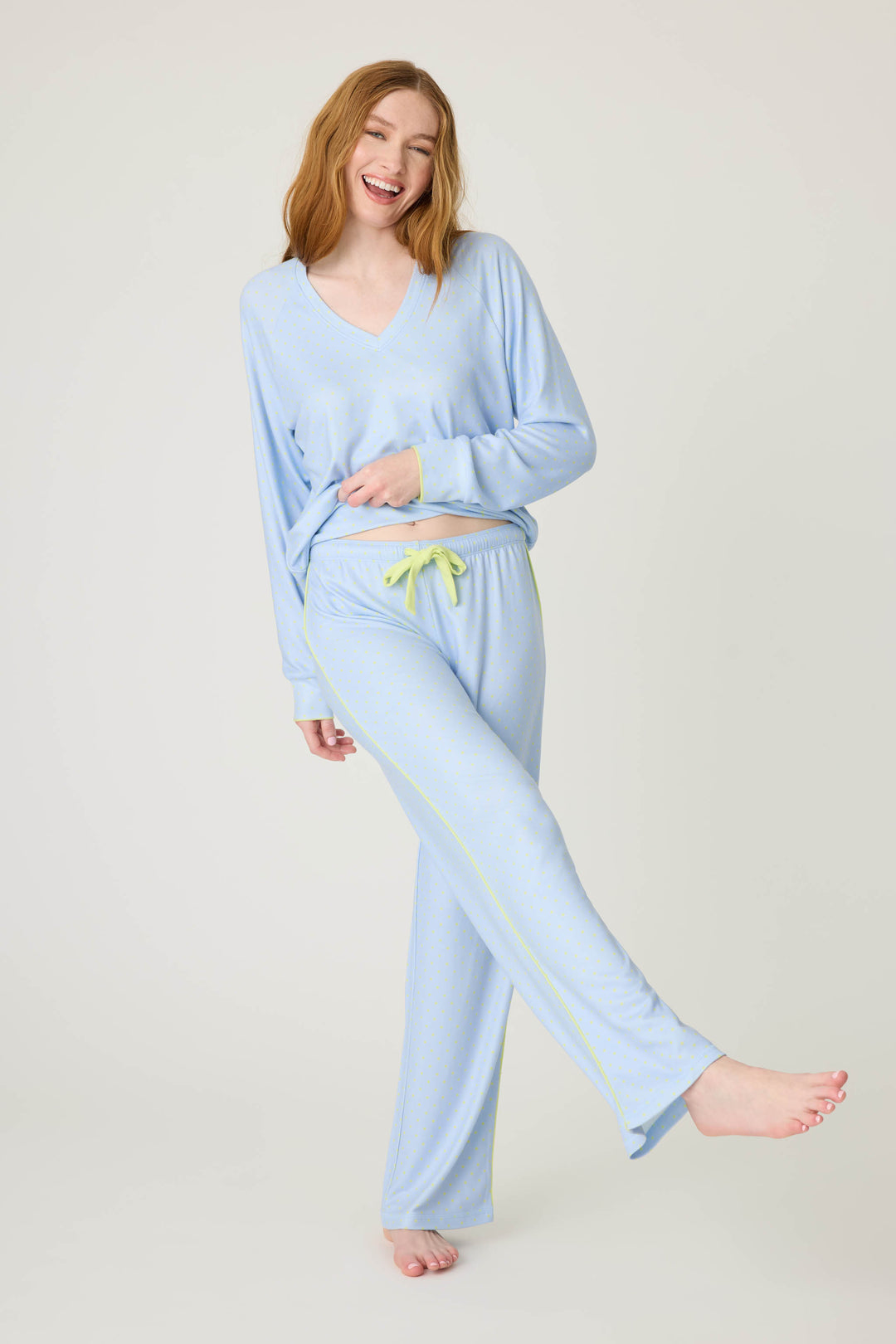 A person with long red hair smiles while wearing the P.J. Salvage Beautiful Life Polka Dot Open Leg Pant pajama set, featuring a v-neck top and matching pants, posing with one leg raised against a plain light background.