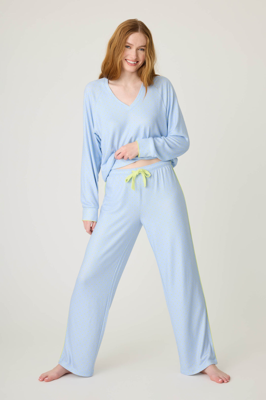 A person with long red hair smiles barefoot against a plain background, wearing the P.J. Salvage Beautiful Life Polka Dot Open Leg Pant and a matching light blue pajama top with a yellow drawstring, holding the hem of the top.