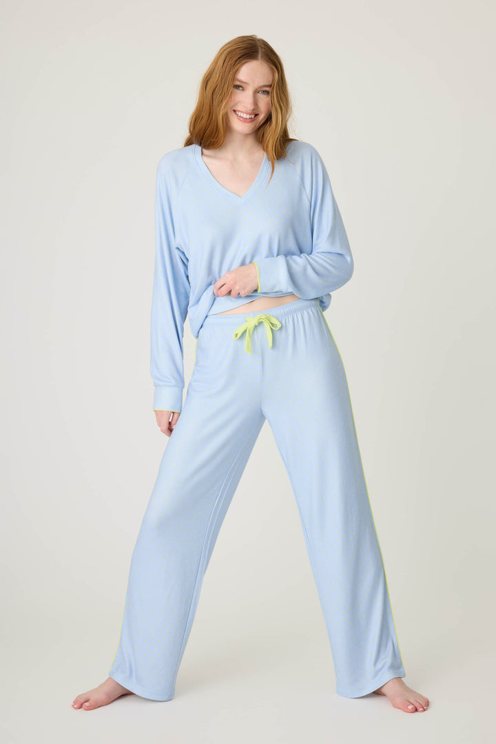 A person with long red hair smiles barefoot against a plain background, wearing the P.J. Salvage Beautiful Life Polka Dot Open Leg Pant and a matching light blue pajama top with a yellow drawstring, holding the hem of the top.