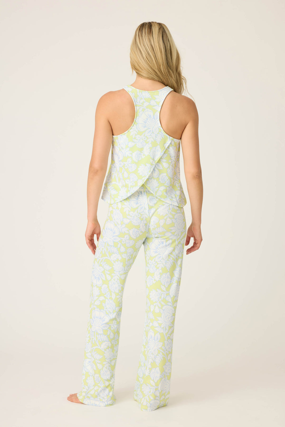 A woman with long blonde hair, shown from the back, models the P.J. Salvage Beautiful Life Floral Print PJ Set— a light green and white sleeveless top with a layered back and matching wide-leg pants—standing barefoot on a plain background.