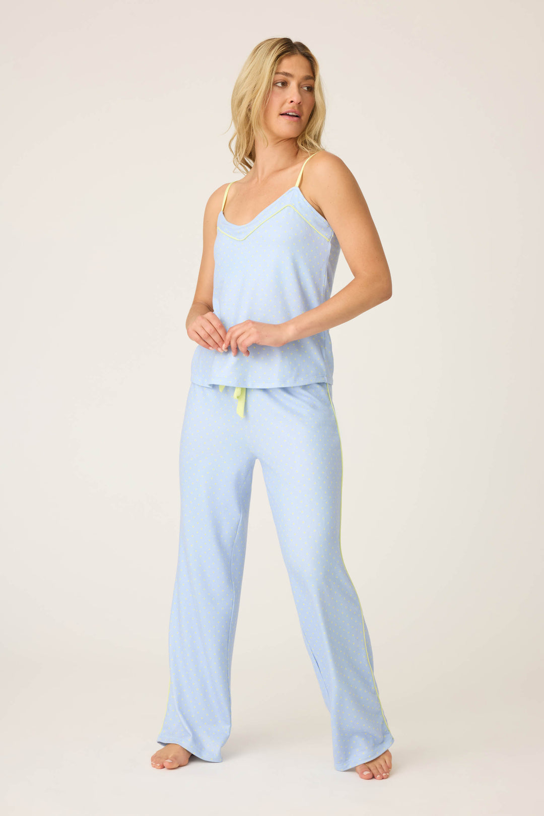 A person stands barefoot in the P.J. Salvage Beautiful Life Polka Dot Pants Set—light blue pajama pants and a matching camisole with thin straps and yellow accents—looking slightly to the side against a plain background.
