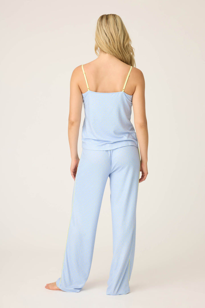 A person with long blonde hair stands barefoot facing away, wearing the P.J. Salvage Beautiful Life Polka Dot Pants Set: a light blue camisole with yellow straps and matching loose pajama pants, against a plain light-colored background.