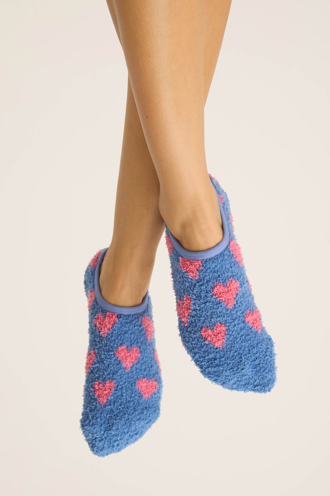 A person with crossed legs wears P.J. Salvage HEARTS SOCKS FLANNELS—blue fuzzy socks with pink hearts—against a plain, light background.