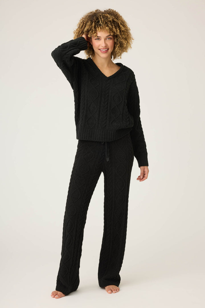 A person with curly hair stands barefoot against a plain background, smiling at the camera in the SOLID PJ SET COZY CHENILLE CABLE KNIT by P.J. Salvage, with one hand touching their hair.