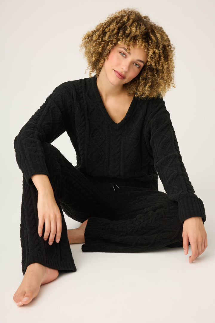 A person with curly hair wears the P.J. Salvage SOLID PJ SET COZY CHENILLE CABLE KNIT in black, sitting on the floor with one leg bent and one extended, looking at the camera against a plain light background.