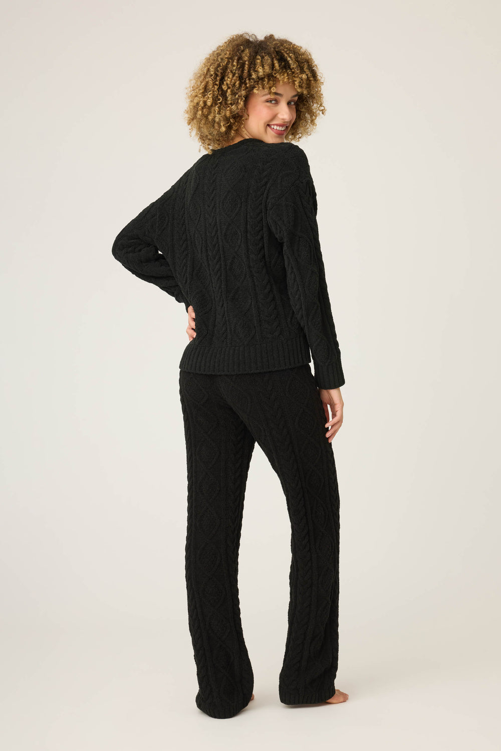 A person with curly hair stands barefoot, facing away and looking over their shoulder, wearing the P.J. Salvage SOLID PJ SET COZY CHENILLE CABLE KNIT against a plain light background.