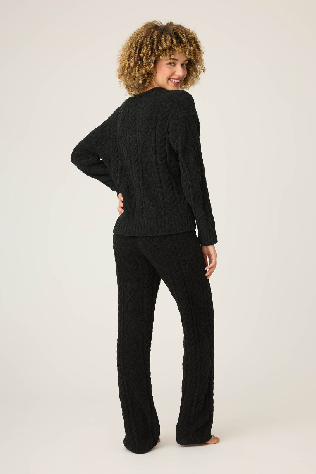 A person with curly hair stands barefoot, facing away and looking over their shoulder, wearing the P.J. Salvage SOLID PJ SET COZY CHENILLE CABLE KNIT against a plain light background.