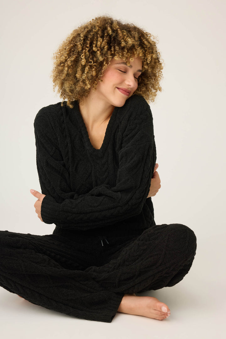 A person with curly hair smiles with eyes closed, sitting cross-legged and hugging themselves, wearing the P.J. Salvage SOLID PJ SET COZY CHENILLE CABLE KNIT against a plain background.