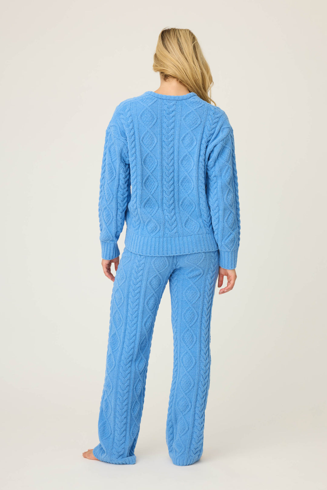 A person with long blonde hair stands barefoot facing away, wearing the P.J. Salvage SOLID PJ SET COZY CHENILLE CABLE KNIT in light blue against a plain background.