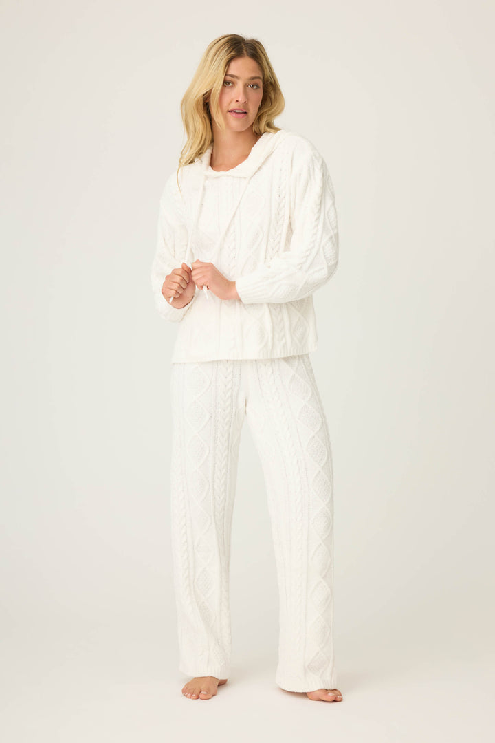 A person stands barefoot against a plain light background, wearing the P.J. Salvage SOLID PJ SET COZY CHENILLE CABLE KNIT—featuring a white textured hoodie and matching knit pants with a diamond pattern.