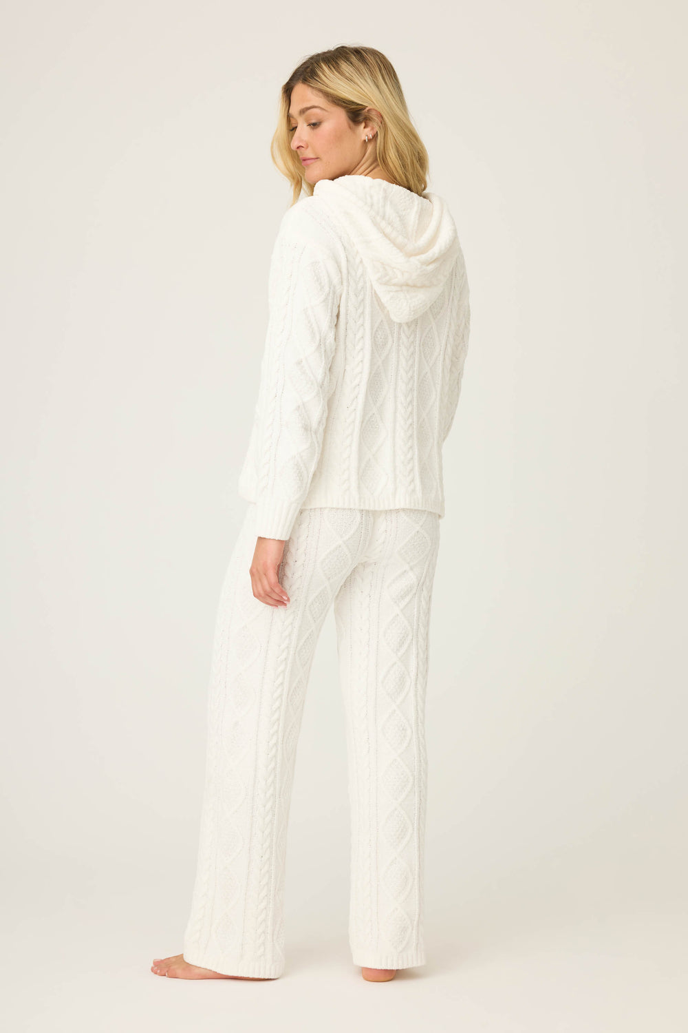 A woman stands barefoot facing away in the SOLID PJ SET COZY CHENILLE CABLE KNIT by P.J. Salvage, featuring a matching white hooded sweater and pants. The background is plain off-white.