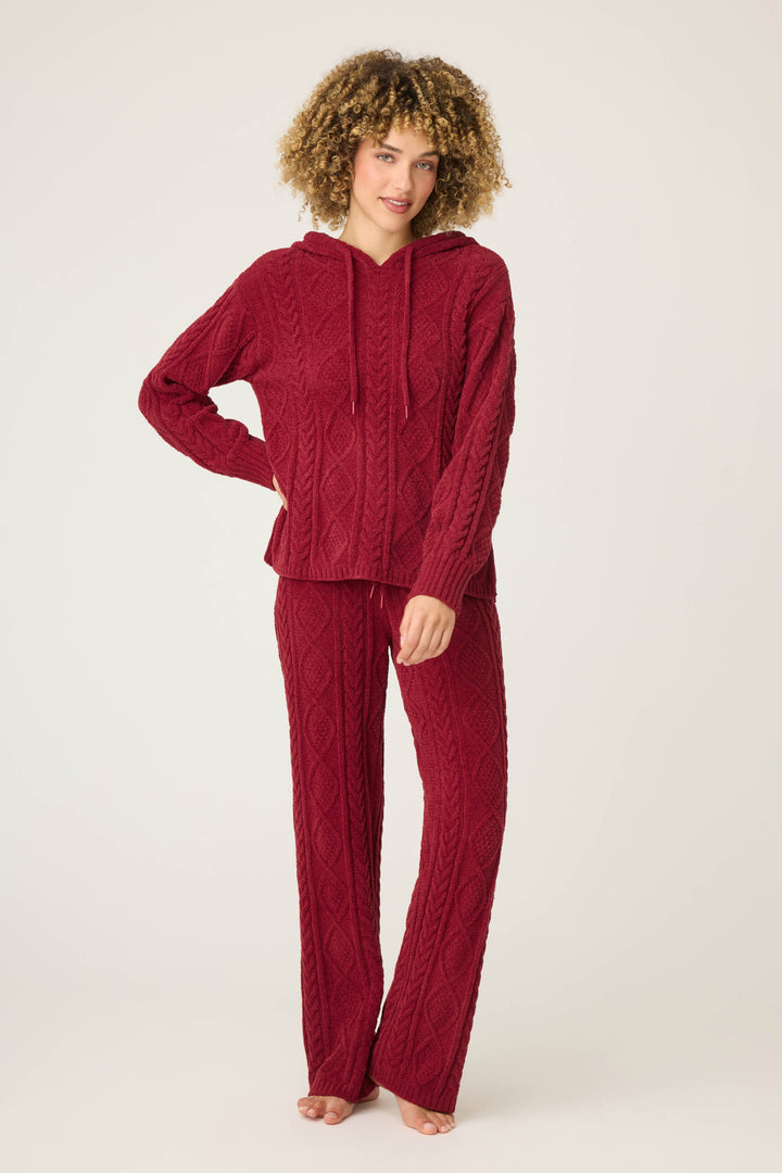 A person stands barefoot against a plain background, wearing the P.J. Salvage SOLID PJ SET COZY CHENILLE CABLE KNIT—a matching textured burgundy hoodie and pants set. Their left hand rests on their hip, and their right hand on their thigh.