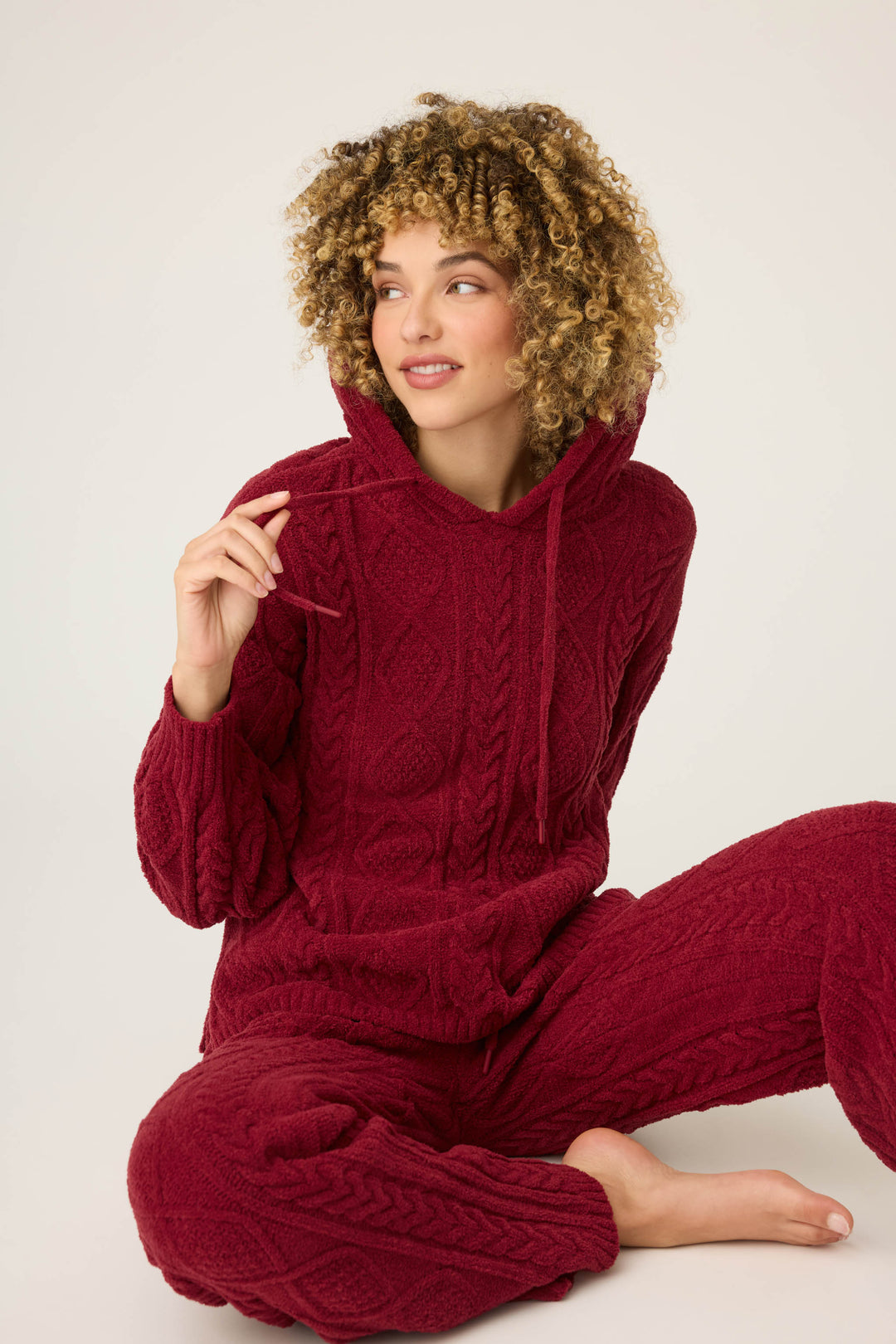 A person with curly blond hair models the P.J. Salvage SOLID PJ SET COZY CHENILLE CABLE KNIT in textured dark red, sitting cross-legged and looking to the side against a plain light background.