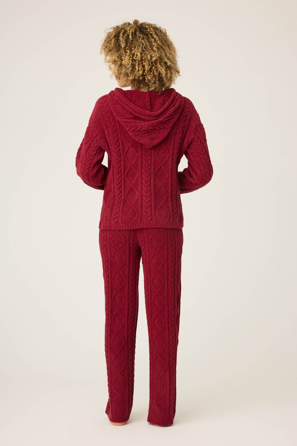A person with curly hair, seen from behind, wears the P.J. Salvage SOLID PJ SET COZY CHENILLE CABLE KNIT in red—a matching textured hoodie and pants—against a plain, light-colored background.