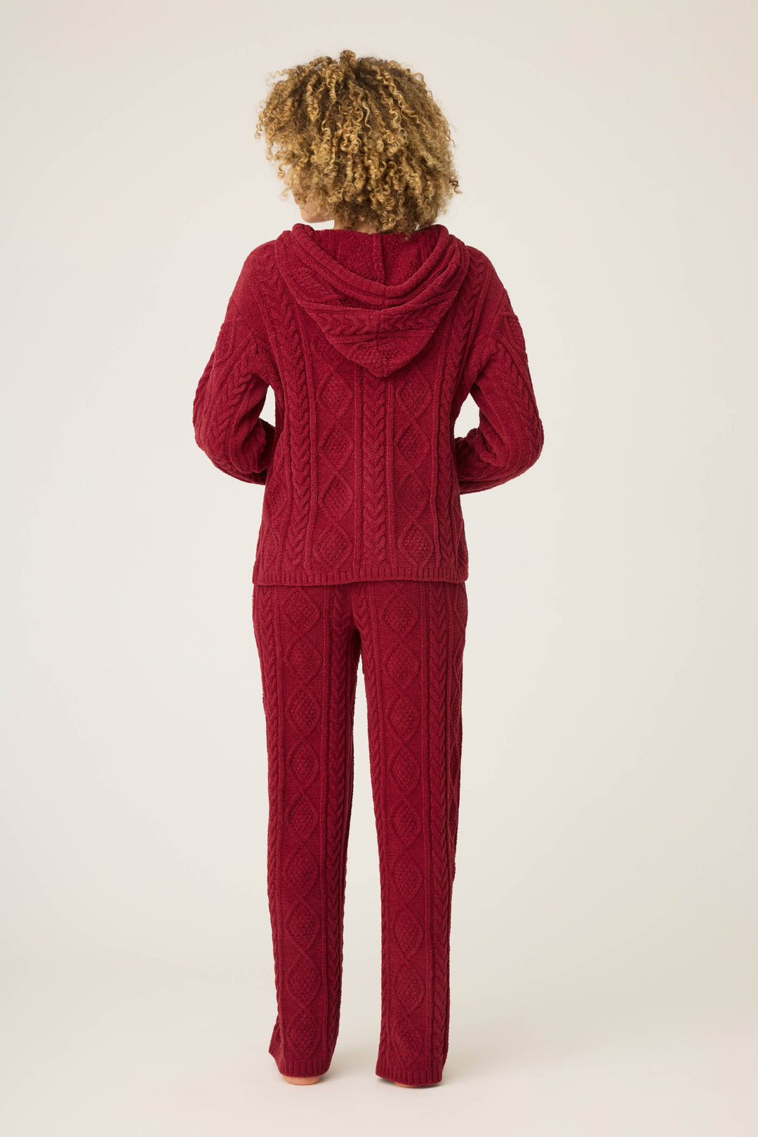 A person with curly hair, seen from behind, wears the P.J. Salvage SOLID PJ SET COZY CHENILLE CABLE KNIT in red—a matching textured hoodie and pants—against a plain, light-colored background.