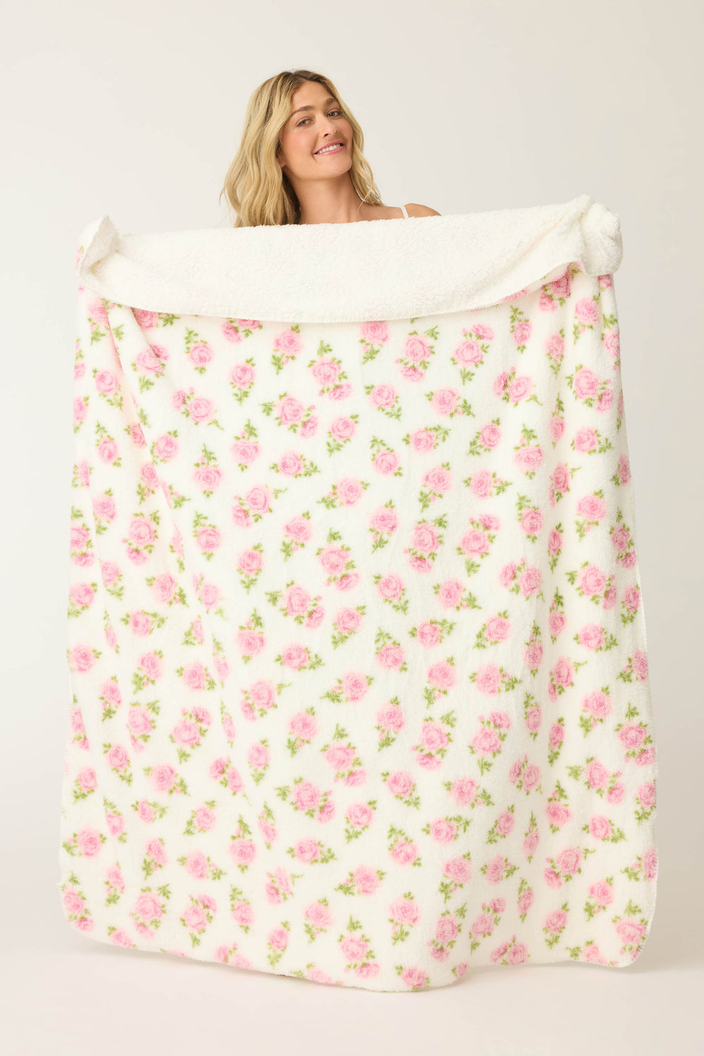 A person with long blonde hair holds up the P.J. Salvage Lets Get Cozy Plush Floral Print Blanket, showcasing its large white surface with a pink and green floral design against a plain, light background.