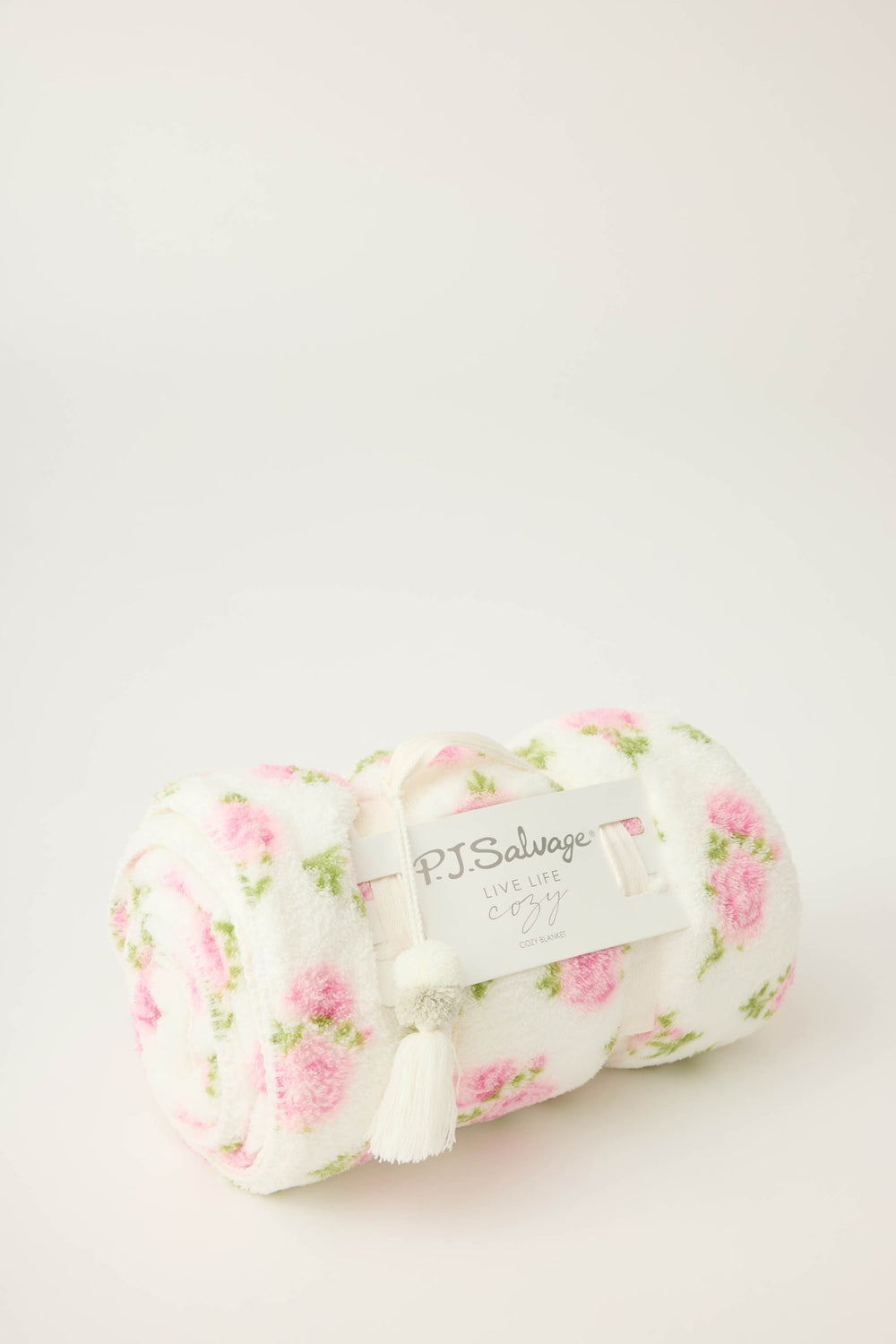 A rolled-up P.J. Salvage Lets Get Cozy Plush Floral Print Blanket features a white base with pink floral designs and green leaves, secured by a label reading Live Life Cozy and adorned with a white tassel. The background is plain and light.