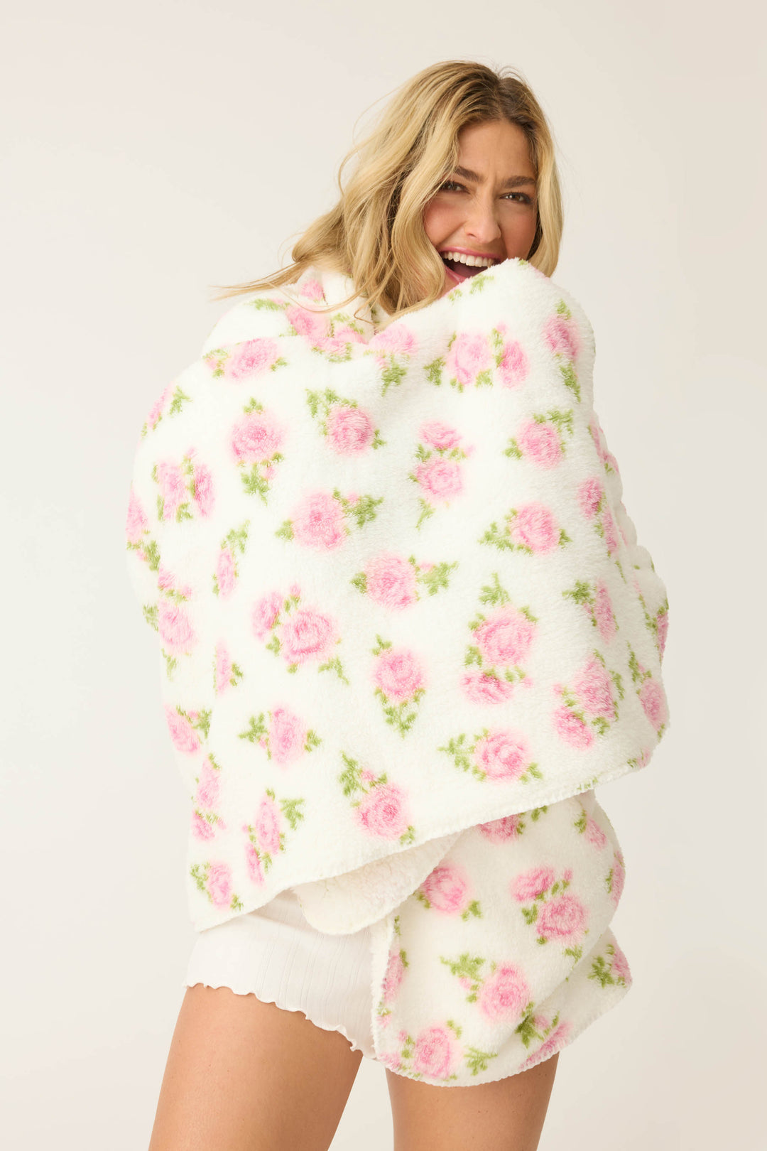 A person with long blonde hair smiles while wrapped in the P.J. Salvage Lets Get Cozy Plush Floral Print Blanket, wearing white shorts and standing against a plain light background.