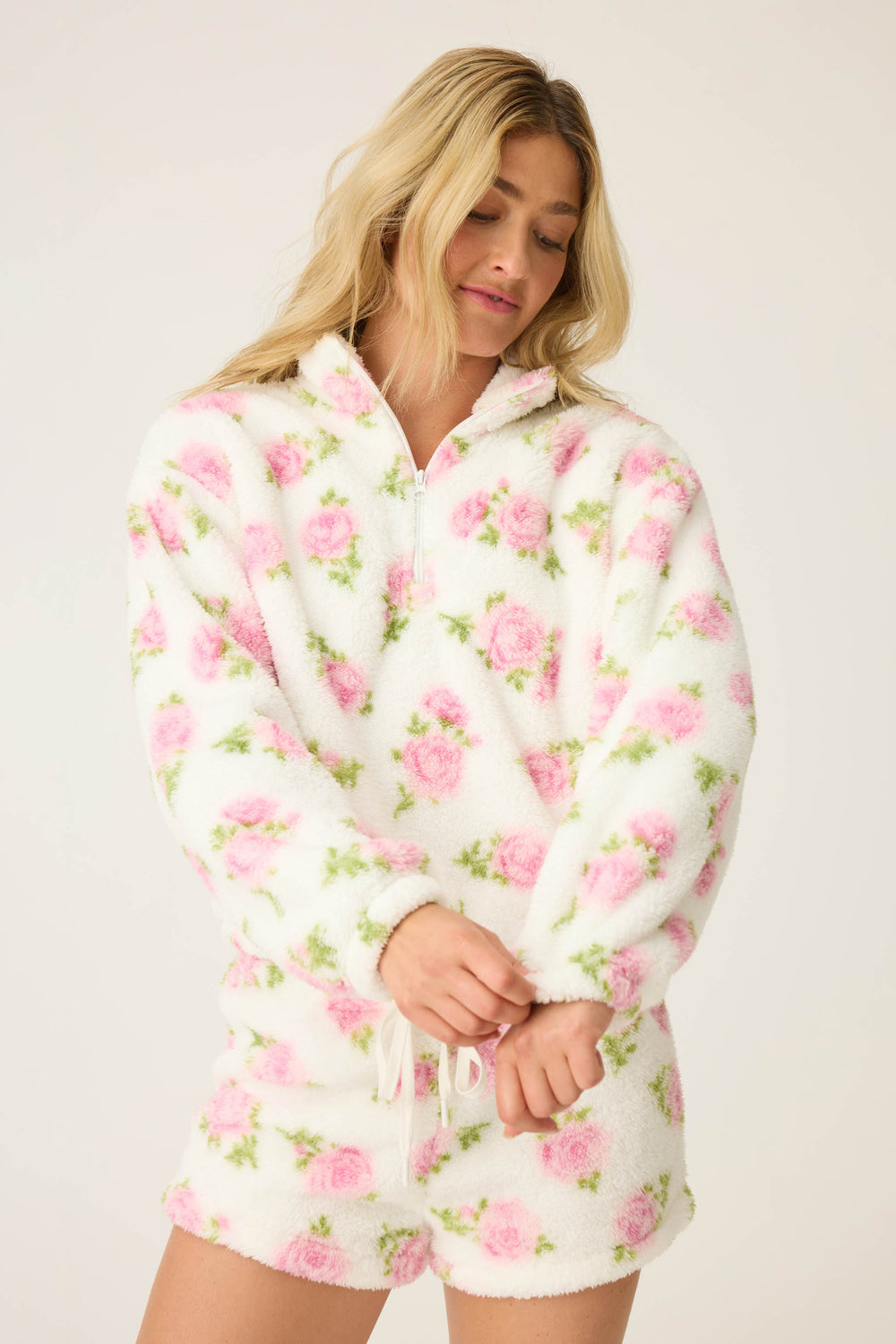 A person with long blonde hair wears the P.J. Salvage Lets Get Cozy Floral Print Jacket, a white fleece pullover with a pink floral pattern, looking down while adjusting the sleeve against a plain light background.