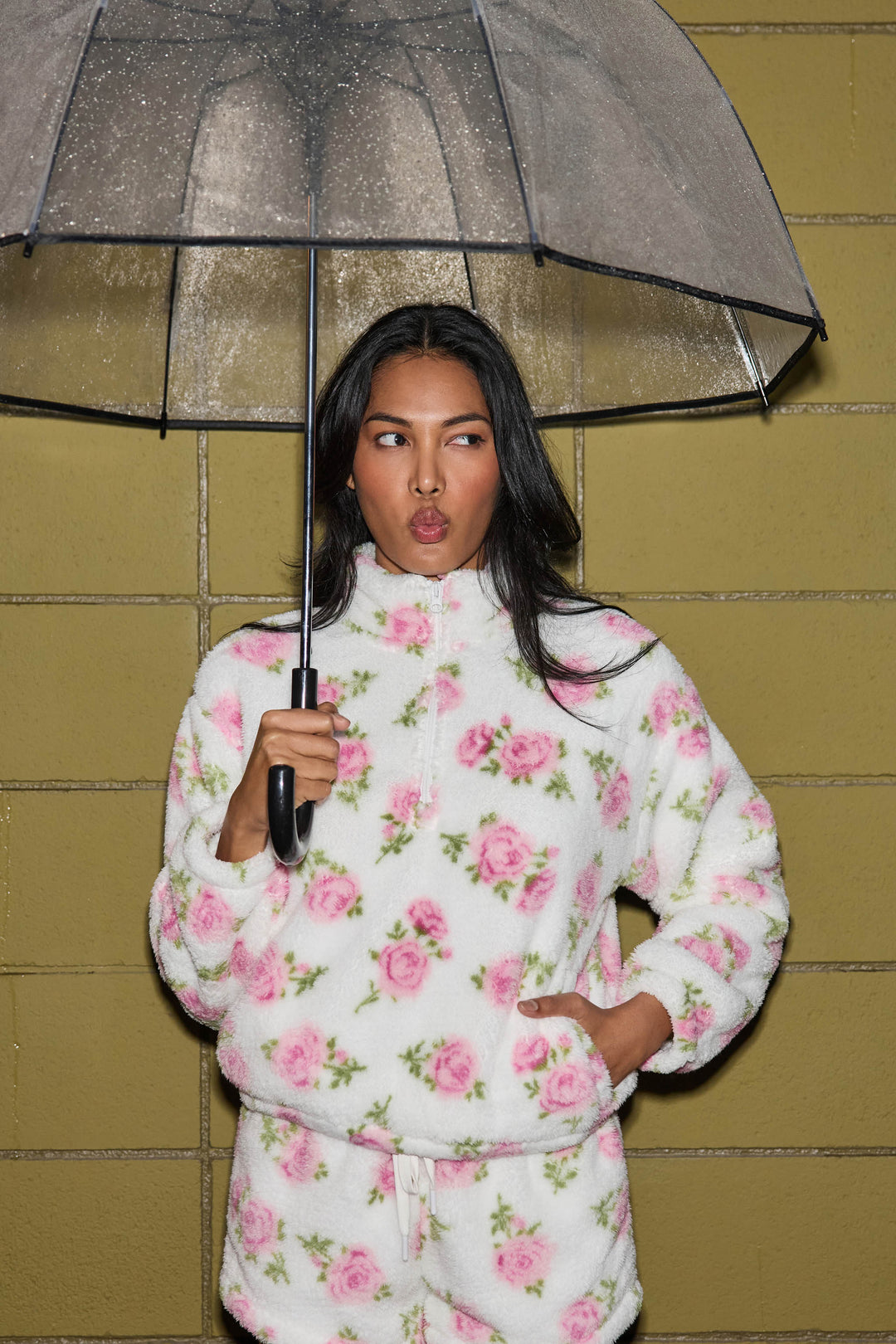 Against a yellow brick wall, a person holds a transparent umbrella and wears the P.J. Salvage Lets Get Cozy Floral Print Jacket, a white fleece with pink flowers. They look thoughtful, gazing slightly to the side.