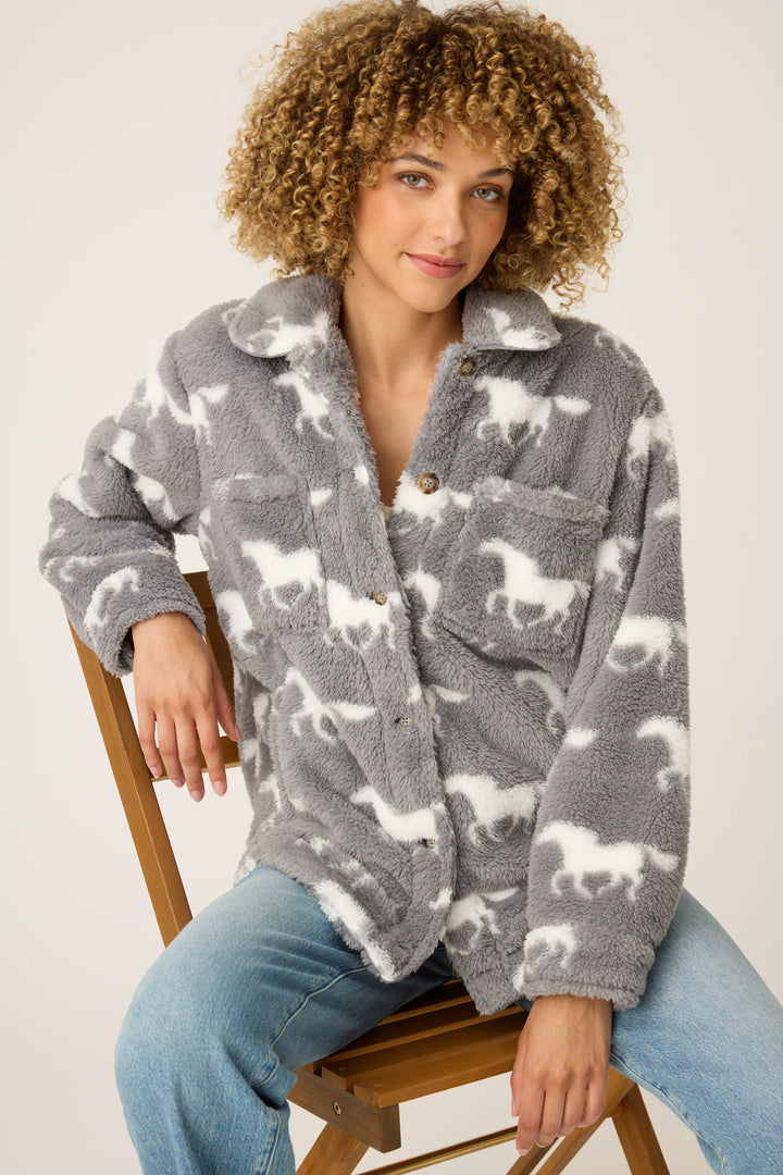 A person with curly hair sits on a wooden chair, wearing the P.J. Salvage HORSES JACKET JACKET LETS GET COZY and light blue jeans, against a plain background.