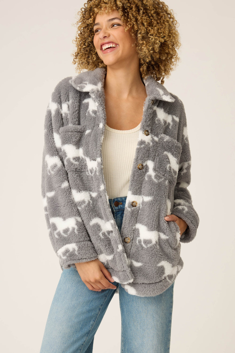 A person with curly hair smiles in the P.J. Salvage HORSES JACKET JACKET LETS GET COZY, a fluffy gray jacket with a white horse pattern, worn over a light ribbed tank top and blue jeans against a plain light background.