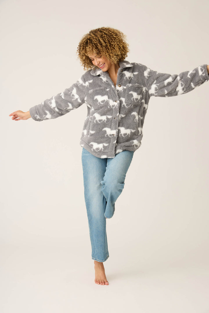 A person with curly hair balances on one bare foot, arms outstretched, wearing the P.J. Salvage HORSES JACKET JACKET LETS GET COZY and blue jeans against a plain light background.