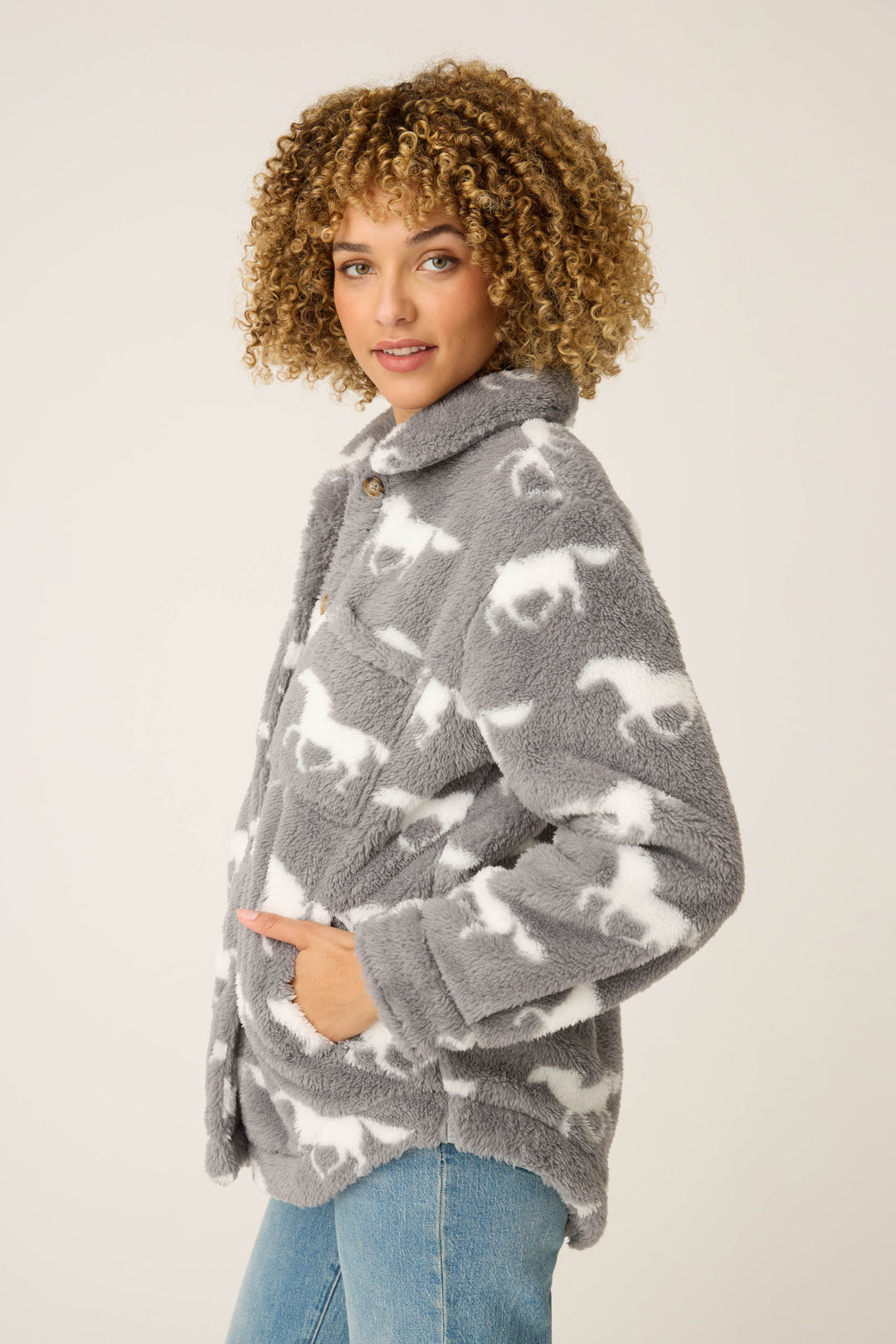 A person with curly hair wears the P.J. Salvage HORSES JACKET JACKET LETS GET COZY, a gray fluffy jacket with a white horse pattern, and blue jeans, standing against a plain light background with one hand in the pocket.