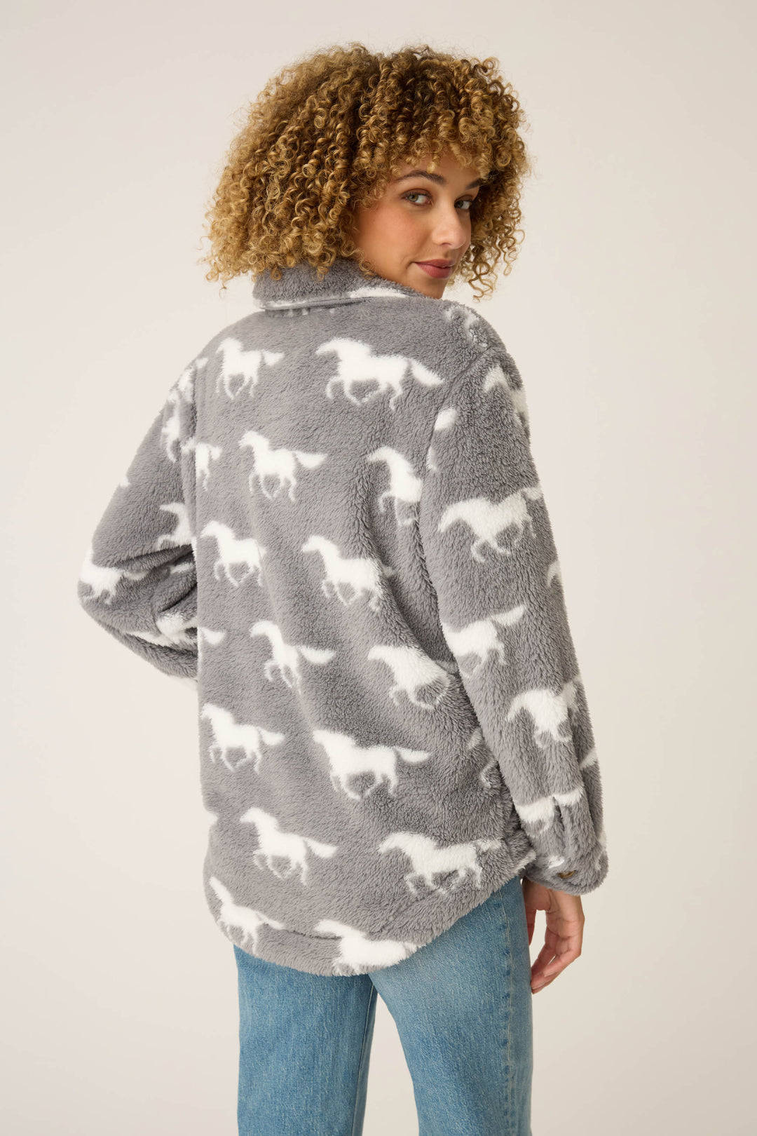 A person with curly hair is facing away, wearing the P.J. Salvage HORSES JACKET JACKET LETS GET COZY— a gray fleece jacket with a white horse pattern— and blue jeans, against a plain light background.
