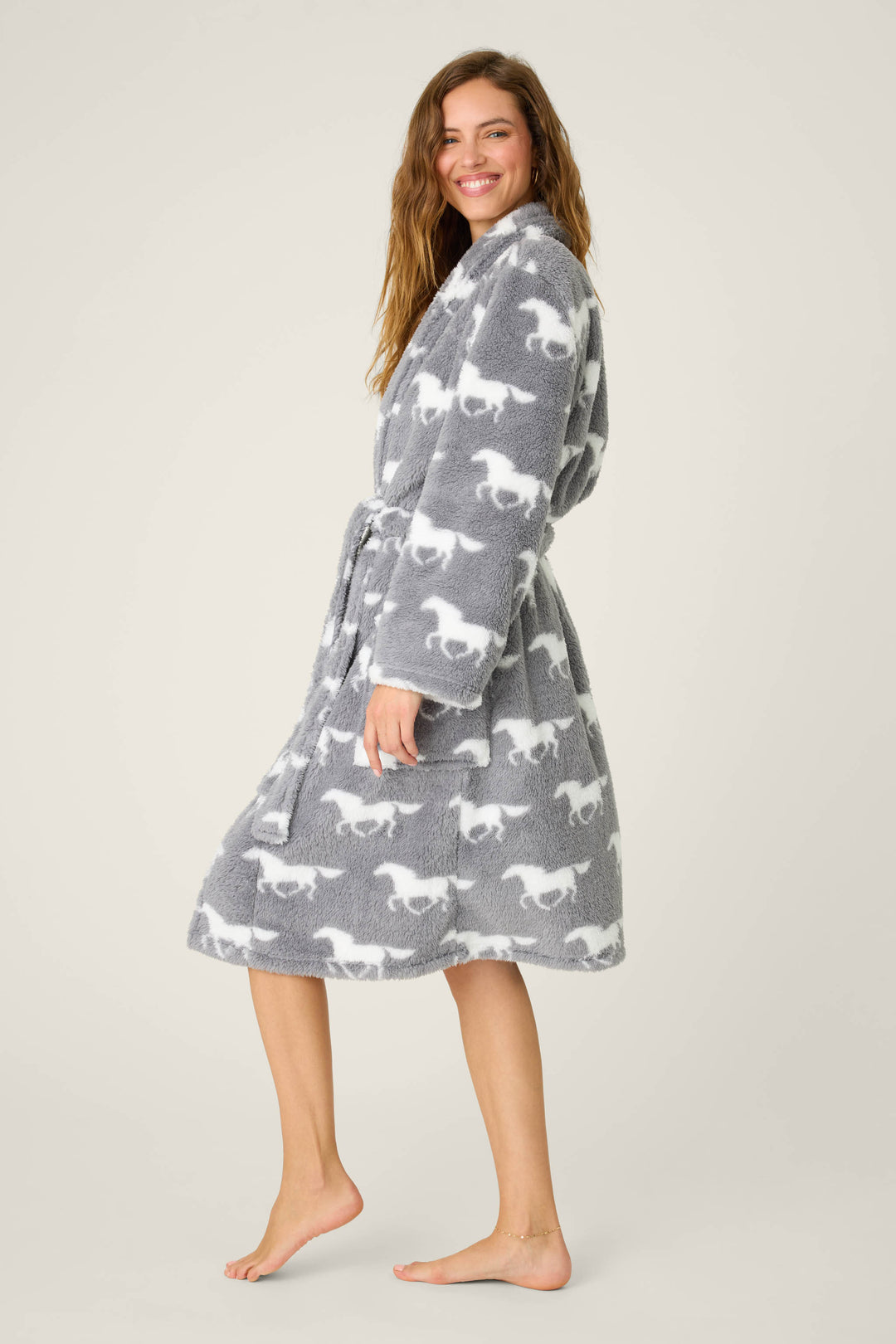 A person stands barefoot against a plain backdrop in the P.J. Salvage STARS ROBE ROBE LETS GET COZY, a grey knee-length robe with long sleeves, pockets, belt, and white dinosaur print. They smile and glance over their shoulder.