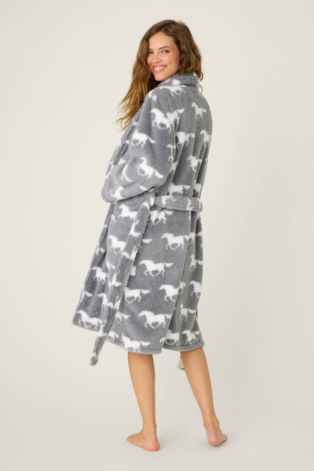 A woman stands barefoot and smiling, wearing the P.J. Salvage STARS ROBE ROBE LETS GET COZY. The cozy grey fleece robe with a belt falls below her knees and features a white horse pattern. She looks over her shoulder, slightly turned to the side.