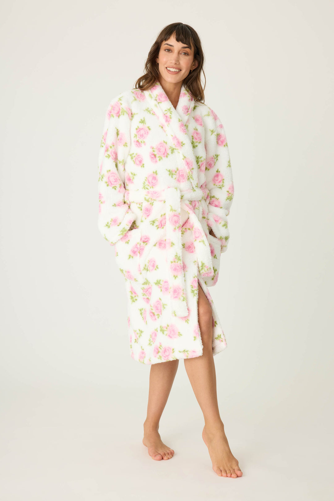 A woman smiles at the camera, barefoot on a plain background, wearing the P.J. Salvage Lets Get Cozy Floral Print Robe with pink flowers, pockets, and a tie belt at the waist.