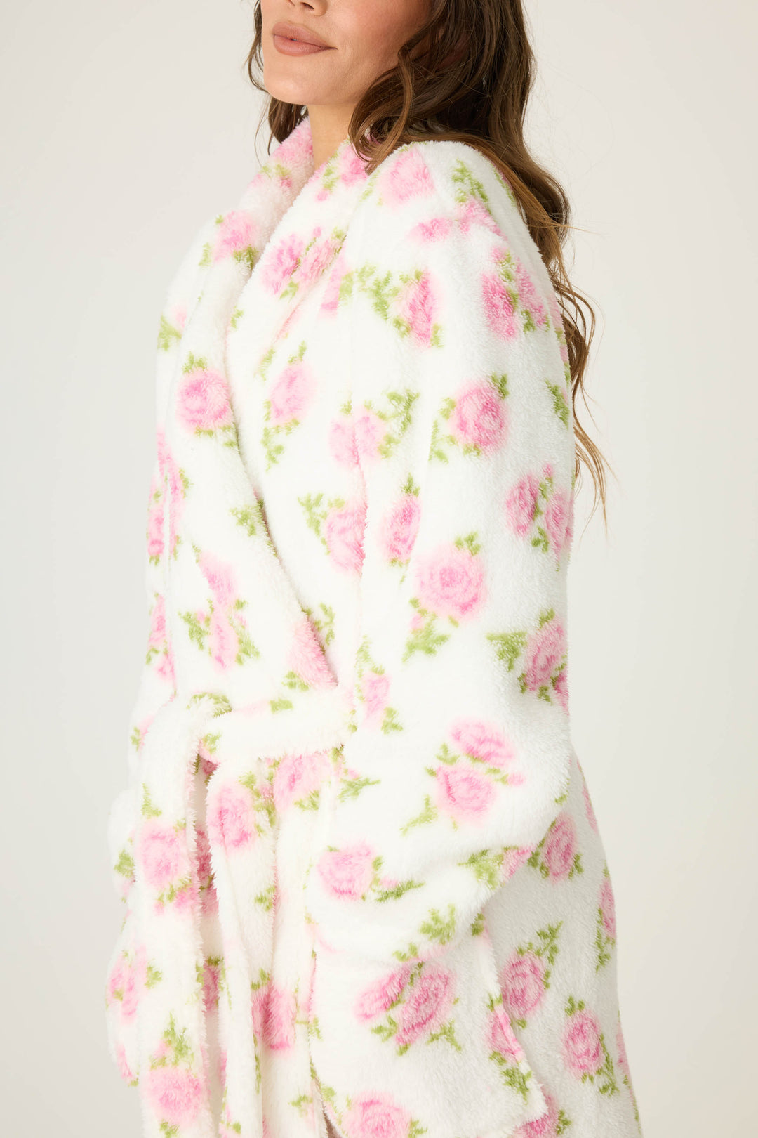A person models the P.J. Salvage Lets Get Cozy Floral Print Robe in white with pink flowers, standing against a plain background. The image is cropped at the shoulders and below the waist, with only part of the face visible.