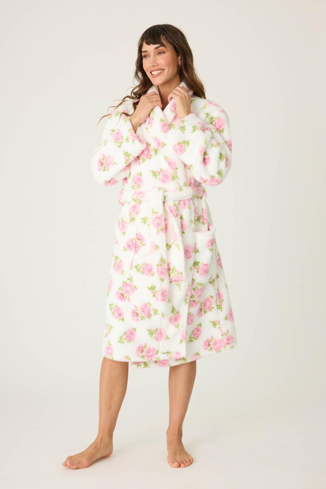 A woman smiles barefoot on a plain background, wearing the P.J. Salvage Let’s Get Cozy Floral Print Robe. She holds the collar of the robe, her long hair loose, showcasing its white fabric with pink floral design.