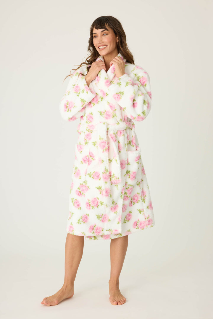 A woman smiles barefoot on a plain background, wearing the P.J. Salvage Let’s Get Cozy Floral Print Robe. She holds the collar of the robe, her long hair loose, showcasing its white fabric with pink floral design.