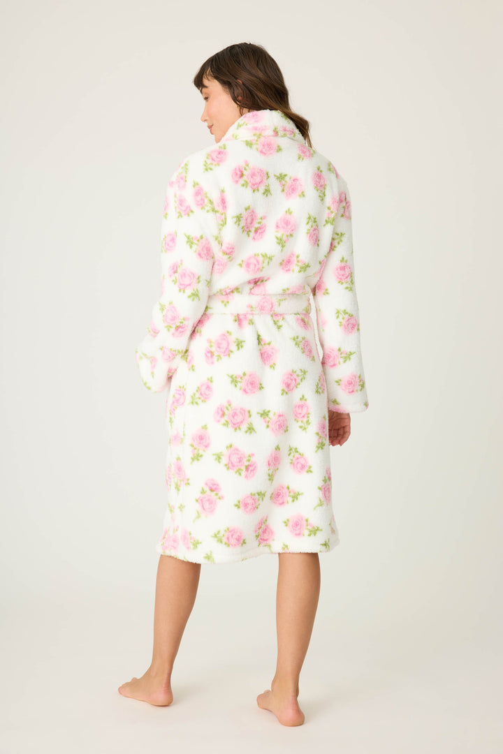 A person stands barefoot facing away, wearing the Lets Get Cozy Floral Print Robe by P.J. Salvage—a white, knee-length robe with a pink floral pattern and waist belt—against a plain, light-colored background.