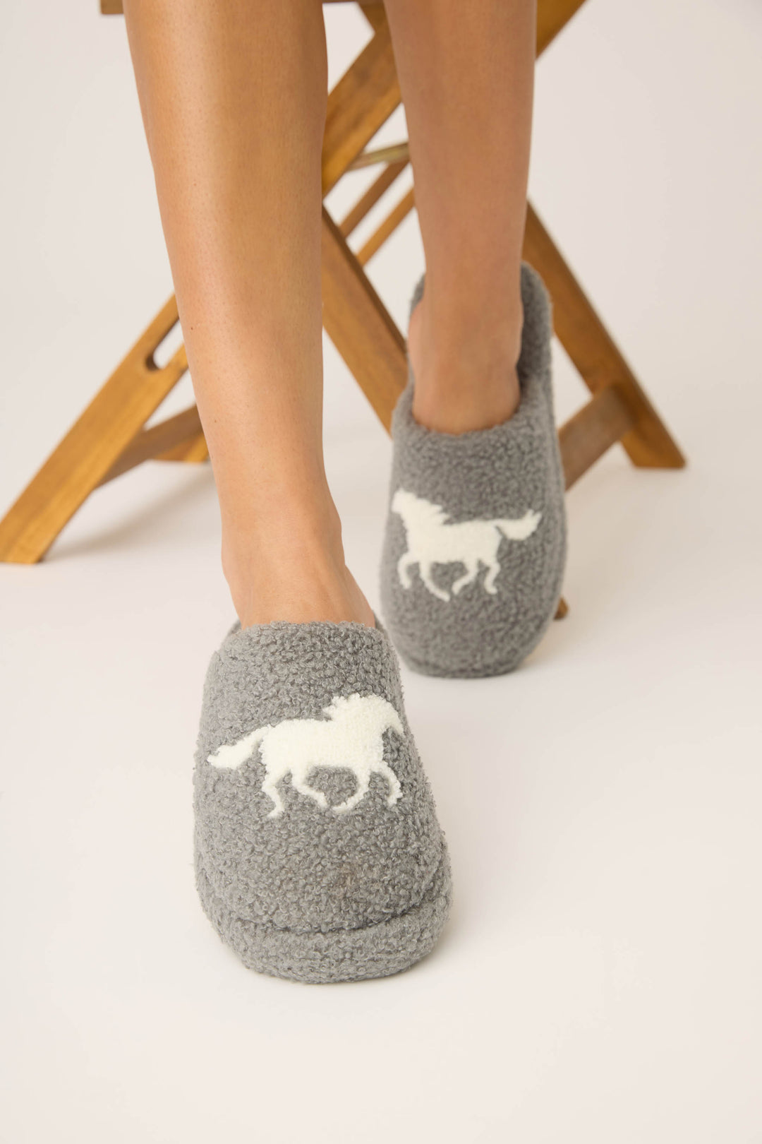 A person wearing P.J. Salvage Let's Get Cozy Horse Fleece Slippers with a galloping horse print stands in front of a wooden stool on a white background.