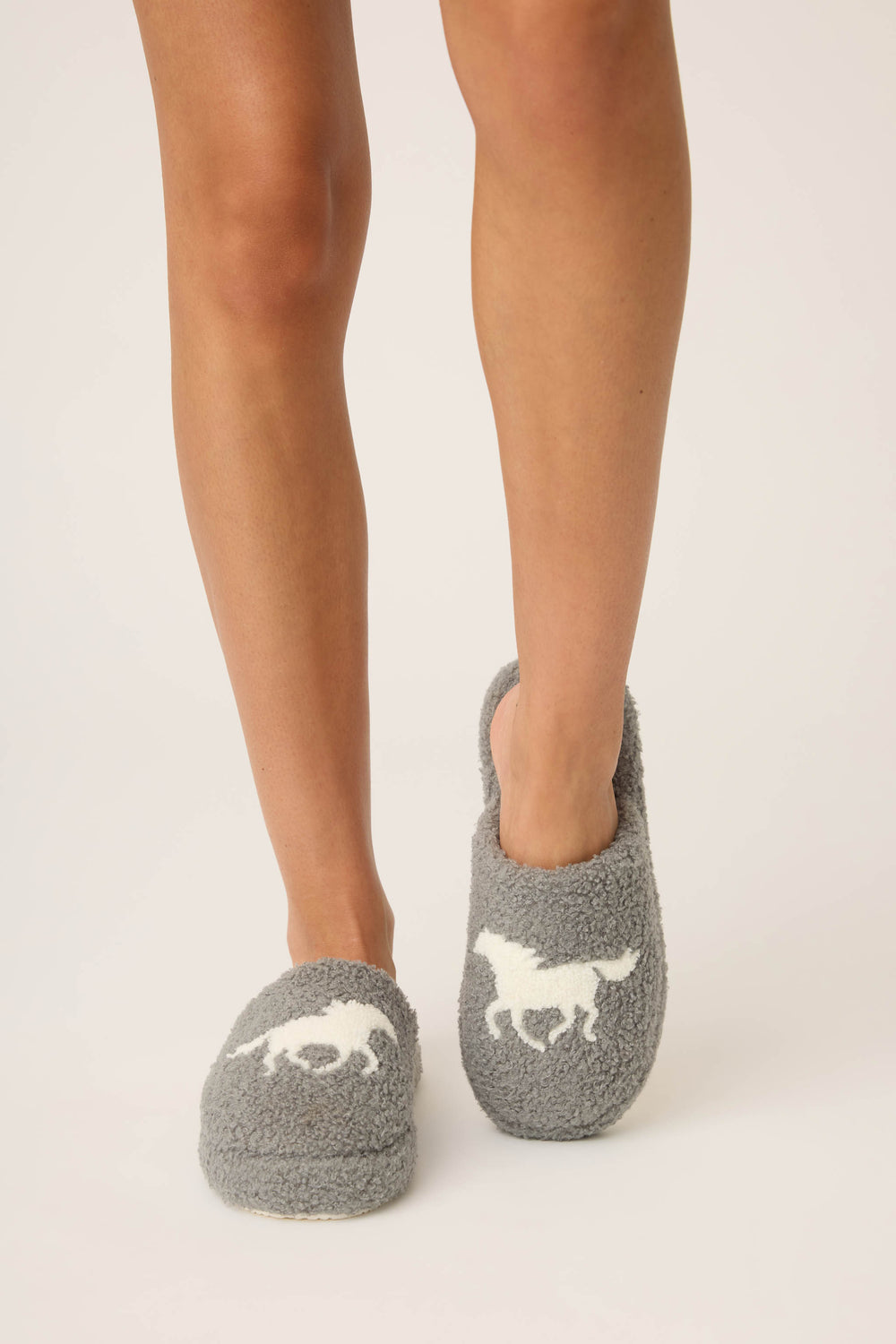 Only the legs and feet are visible as a person stands on a white background, wearing P.J. Salvage Let’s Get Cozy Horse Fleece Slippers featuring fuzzy gray fabric and a galloping horse print on top.