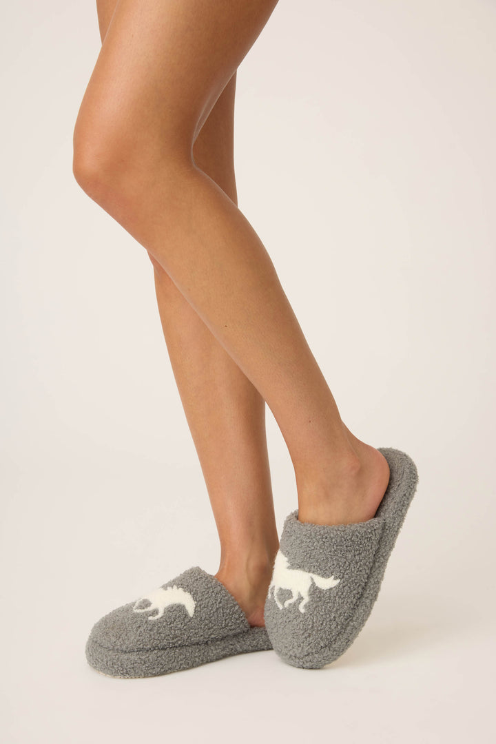 A person stands on tiptoe wearing P.J. Salvage’s Let’s Get Cozy Horse Fleece Slippers—gray and fuzzy, each featuring a white horse silhouette—against a plain, light-colored background.