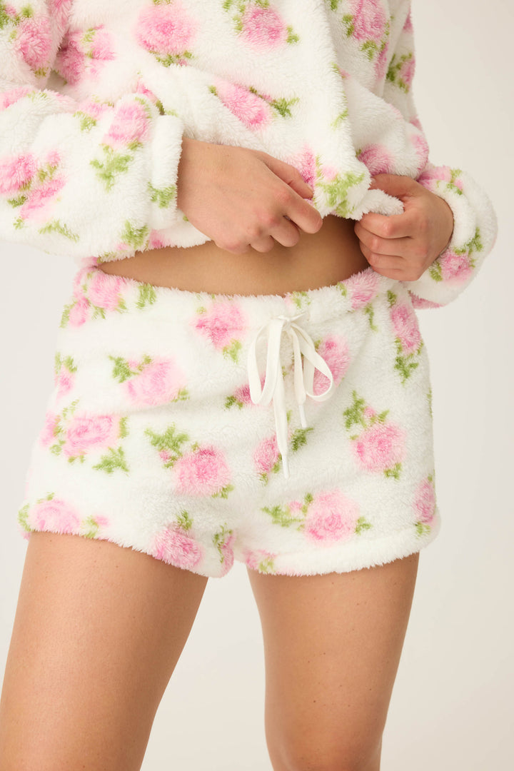 A person wears P.J. Salvage Let’s Get Cozy Floral Print Shorts, shown from torso to legs and adjusting the waistband; the matching fuzzy pajama set features a pink and green floral pattern.