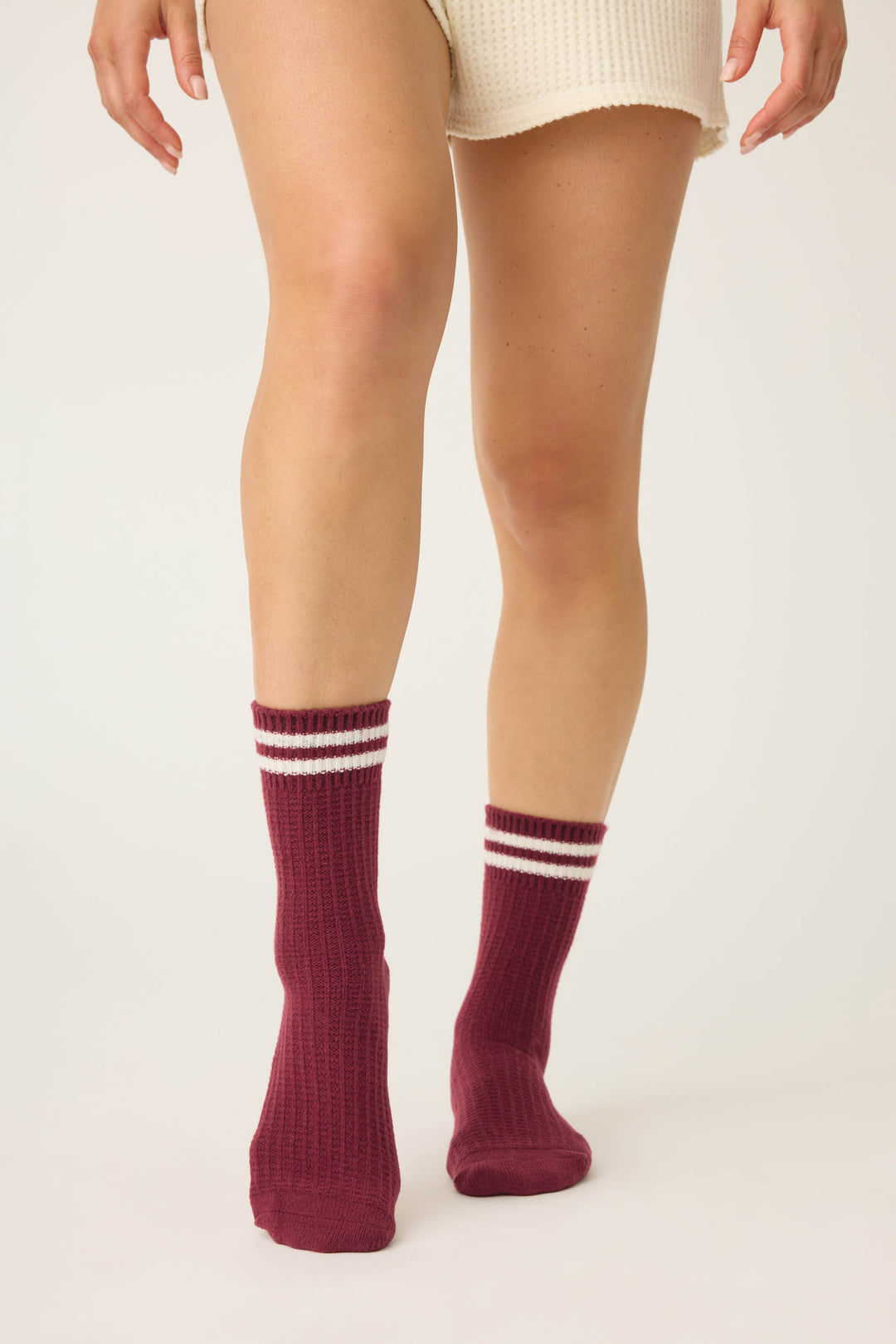 Only the legs and lower torso are visible of a person standing on a light surface, wearing cream-colored shorts and maroon P.J. Salvage SOLID SOCKS LETS GET COZY with two white stripes near the top.