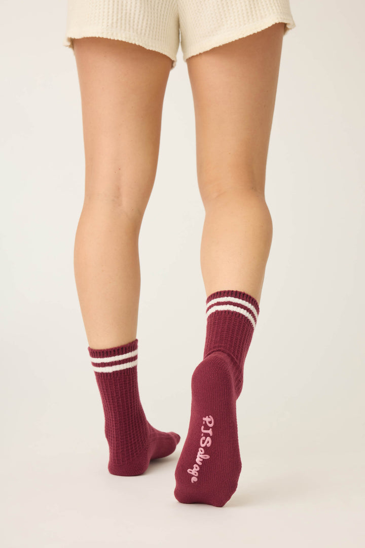 A person stands with their back to the camera, wearing cream shorts and P.J. Salvage SOLID SOCKS LETS GET COZY—maroon socks with two white stripes and “P.S. Say hey” in white on the bottom of one sock.
