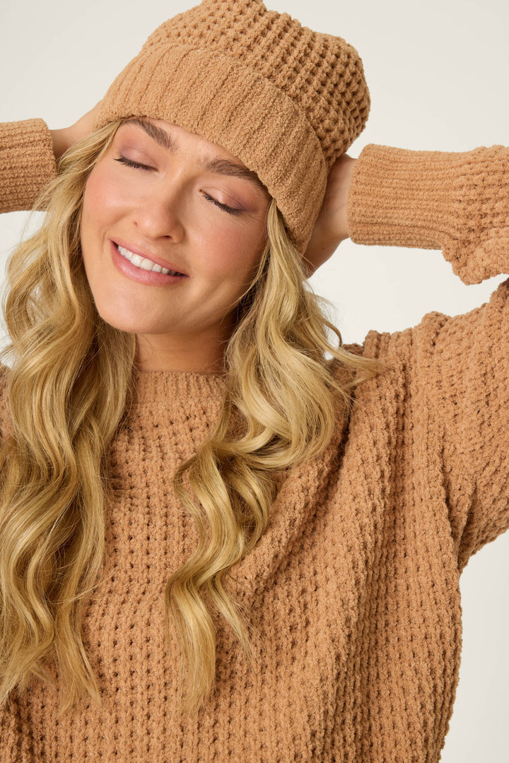 A person with long blonde hair smiles with eyes closed, wearing a textured tan sweater and the P.J. Salvage Chenille Waffle Caramel Beanie, with hands resting behind their head.