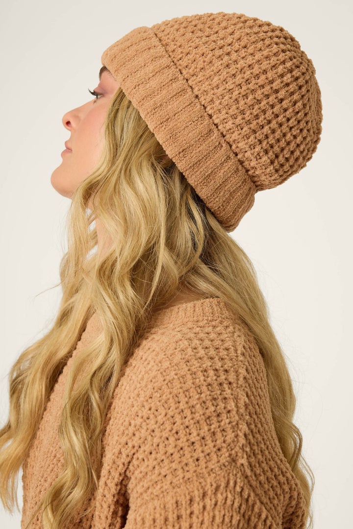 A person with long, wavy blonde hair is shown in profile wearing the P.J. Salvage Chenille Waffle Caramel Beanie and a matching sweater against a plain light background.