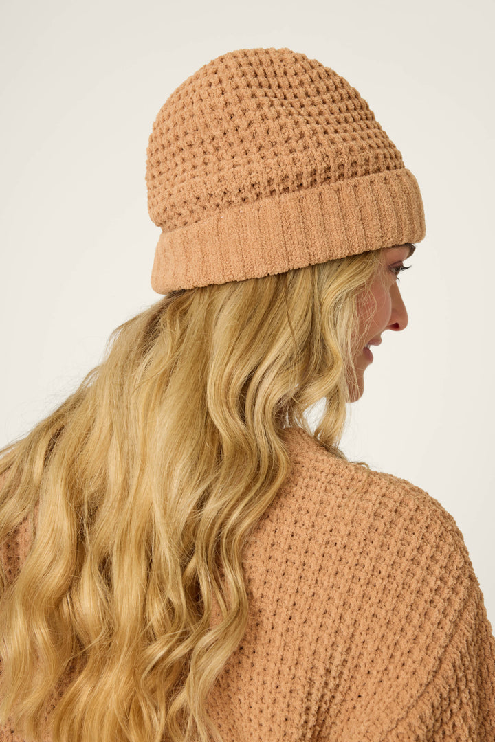 A person with long, wavy blonde hair is seen from the side and back, wearing the P.J. Salvage Chenille Waffle Caramel Beanie with a matching sweater against a plain, light background.