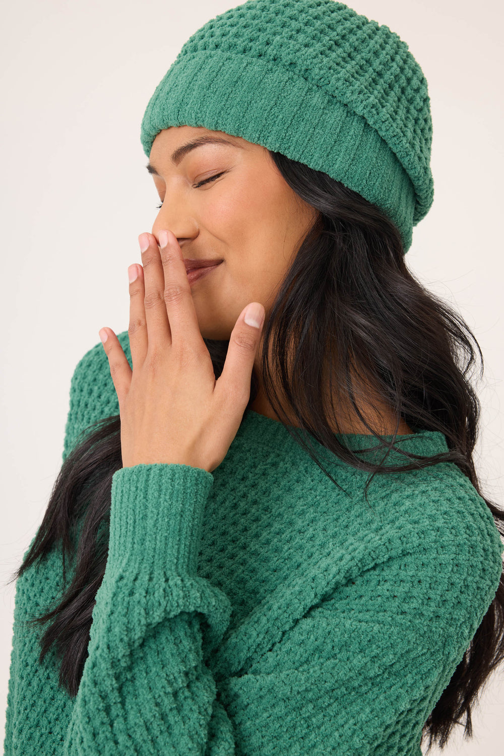 A person with long dark hair smiles with closed eyes, wearing the P.J. Salvage Chenille Waffle Forest Beanie and a matching sweater, holding their hand near their mouth against a plain background.