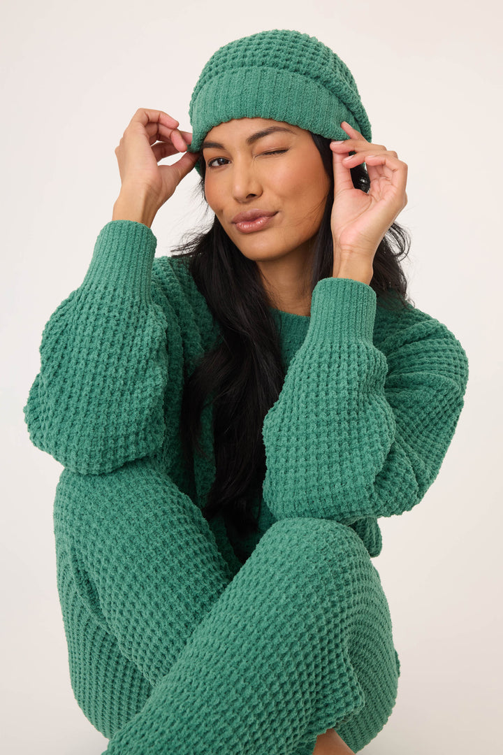 Wearing the P.J. Salvage Chenille Waffle Forest Beanie, a person in matching green knit attire sits with one knee up, winking and adjusting the beanie with both hands against a plain light background.