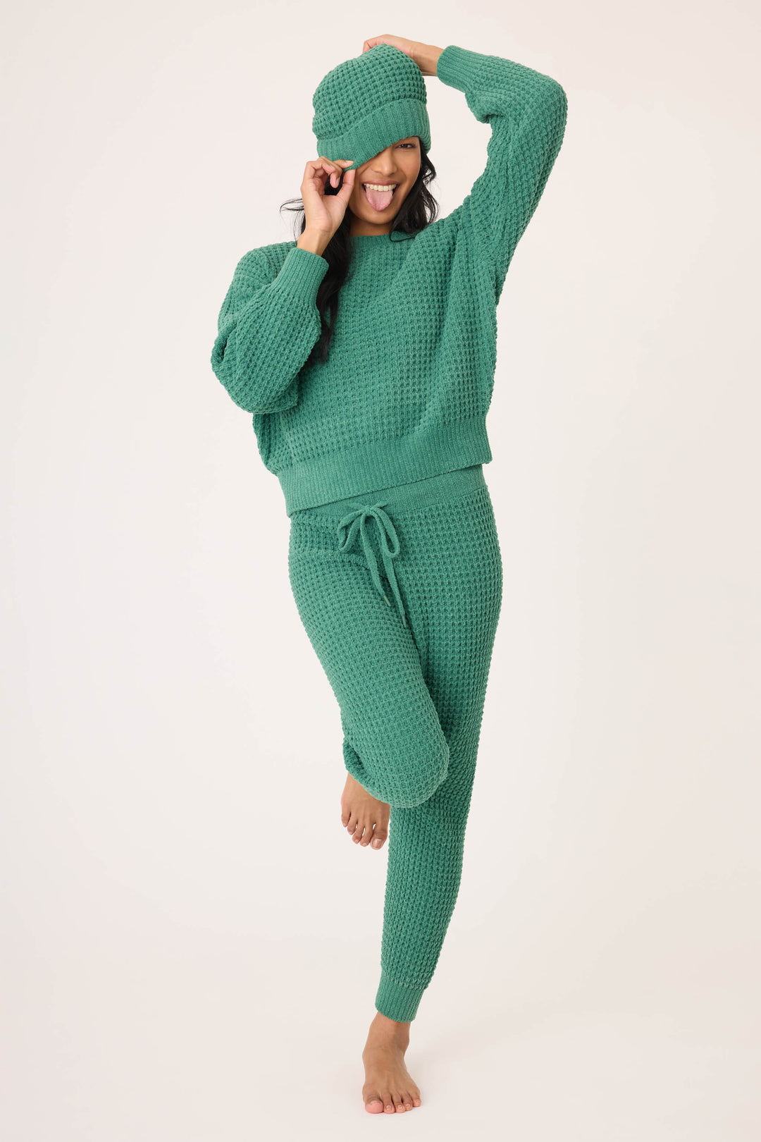 A person stands barefoot on one leg, smiling, in a matching green knit sweater, joggers, and the Chenille Waffle Forest Beanie by P.J. Salvage. The background is plain and light.