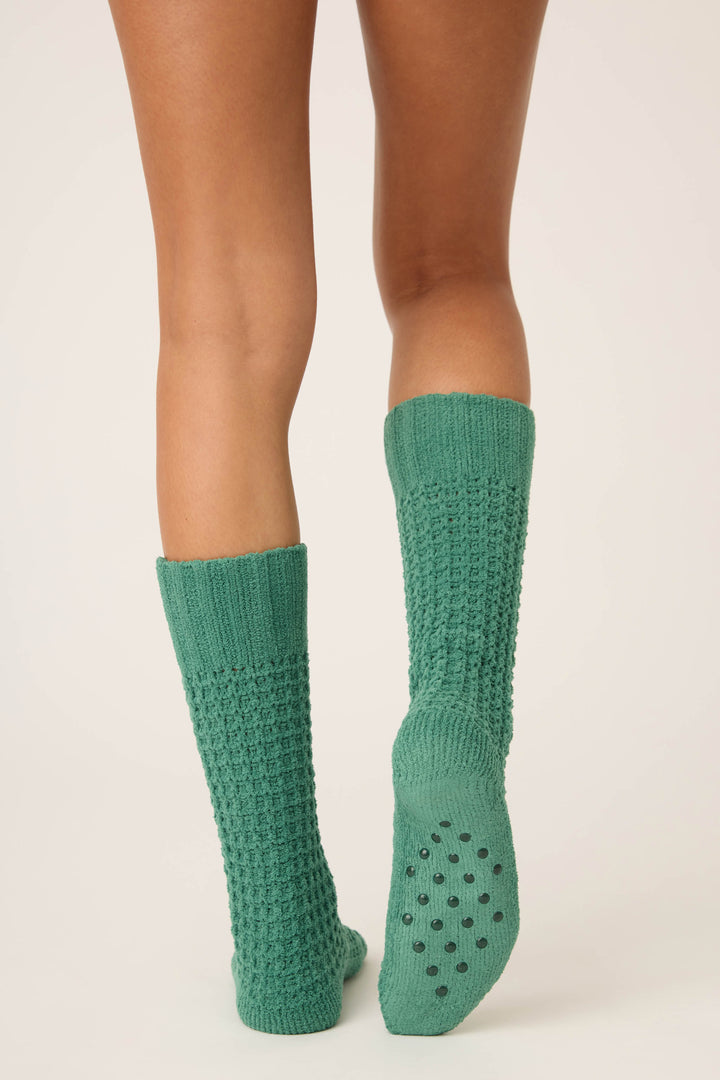 A person stands with legs shown from the knees down, wearing P.J. Salvage Chenille Waffle Solid Crew Socks in textured green. The left sock’s sole features dark grip dots. Background is plain and light-colored.