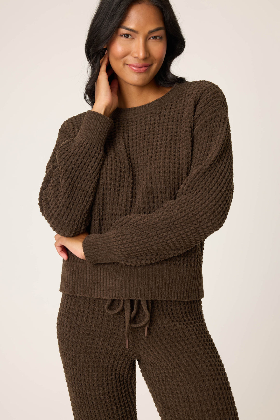A person with long, dark hair wears the P.J. Salvage Chenille Waffle Solid Chocolate Sweater, smiling gently with one hand touching their face and the other arm crossed in front of them.
